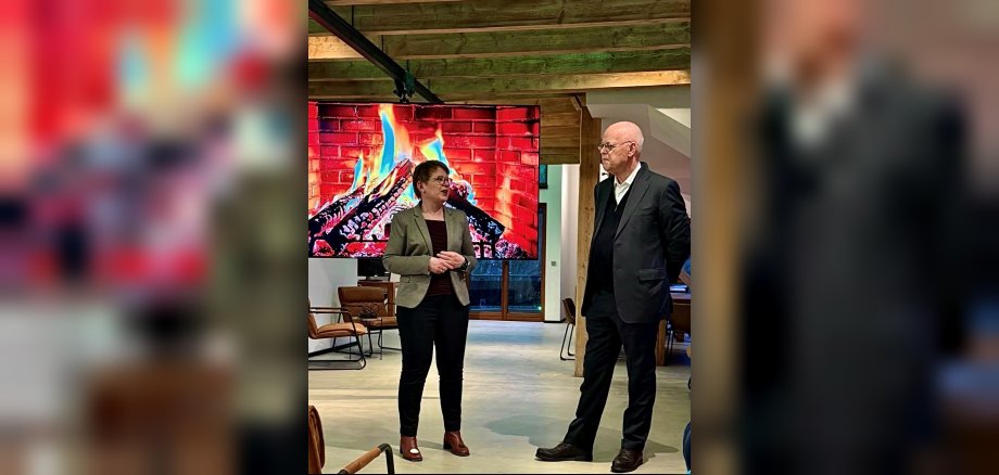 Dr. Tanja Machalet und Rudolph Scharping vor einer Leinwand, die ein brennendes Kaminfeuer zeigt