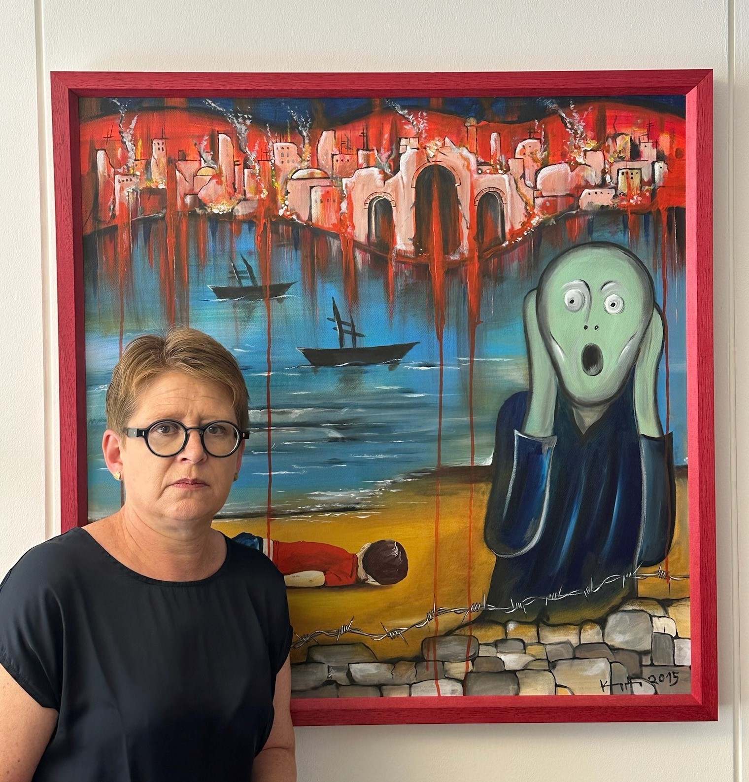 Dr. Tanja Machalet vor dem Gemälde des Nauorter Künstlers Heinz Kuppler. Das Gemälde ist an das bekannte Werk von Edvard Munch "Der Schrei" angelehnt. Im Vordergrund eine stilisierte Person, die mit weit aufgerissenem Mund zu schreien scheint, im Hintergrund in sehr kräftigen Farben eine brennende Stadt, weiter vorne das Meer mit zwei Booten, die nur aus Schatten zu bestehen scheinen. Ganz im Vordergrund ein Kinderkörper am Strand in Erinnerung an Alan Kurdi, der auf der Flucht ertrunken ist und dessen Foto um die Welt ging.