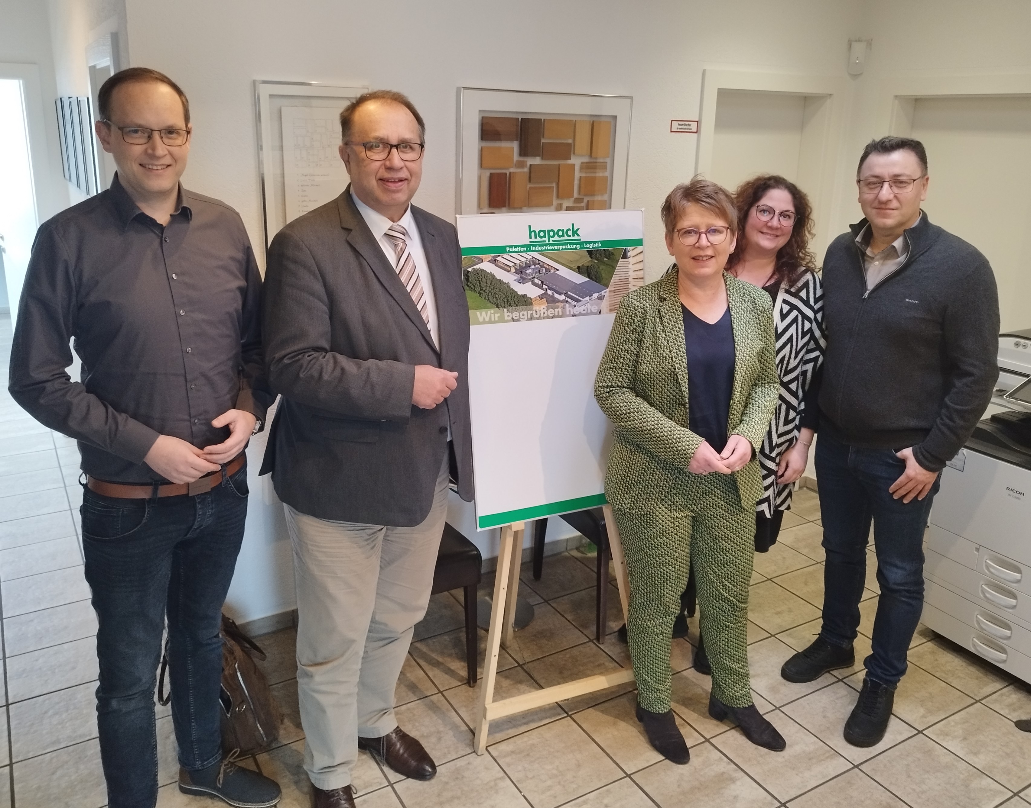 Das Foto zeigt von links: Clint Sikorski, Richard Hover, Dr. Tanja Machalet, Laura Heuchemer und Yüksel Ergün vor einer Tafel mit rechteckigen Materialmustern aus Holz in verschiedenen Farben, die an der Wand hängt. Ebenfalls im Hintergrund steht auf einer Staffelei ein Foto mit einer Luftaufnahme vom Firmengebäude der Fa. hapack.