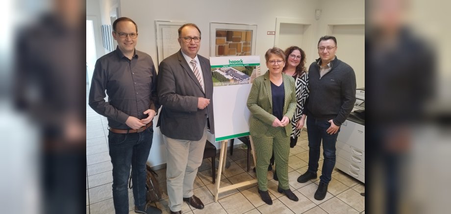 Das Foto zeigt von links: Clint Sikorski, Richard Hover, Dr. Tanja Machalet, Laura Heuchemer und Yüksel Ergün vor einer Tafel mit rechteckigen Materialmustern aus Holz in verschiedenen Farben, die an der Wand hängt. Ebenfalls im Hintergrund steht auf einer Staffelei ein Foto mit einer Luftaufnahme vom Firmengebäude der Fa. hapack.