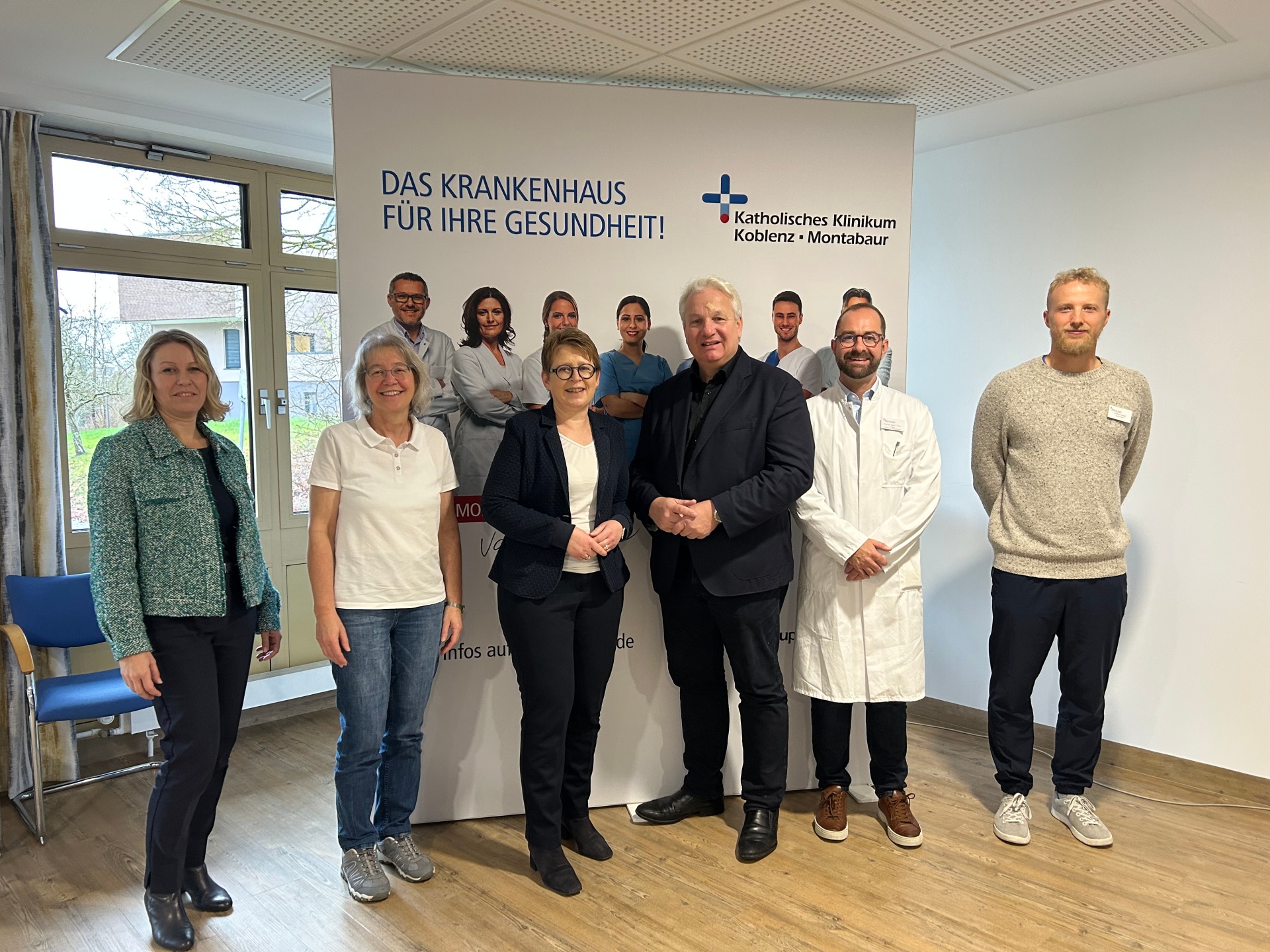 Im Bild von links nach rechts stehend in einem hellen Raum mit Holzboden und hellen Wänden, im Hintergrund ein bodenlanges Fenster, teilweise vor dem Fenster ein Roll-up mit Werbung vom Klinikum: Fr. Saal, Fr. Dr. Maxeiner, Fr. Dr. Machalet, Hr. Isenberg, Hr. Prof. Dr. Fink, Hr. Heuser
