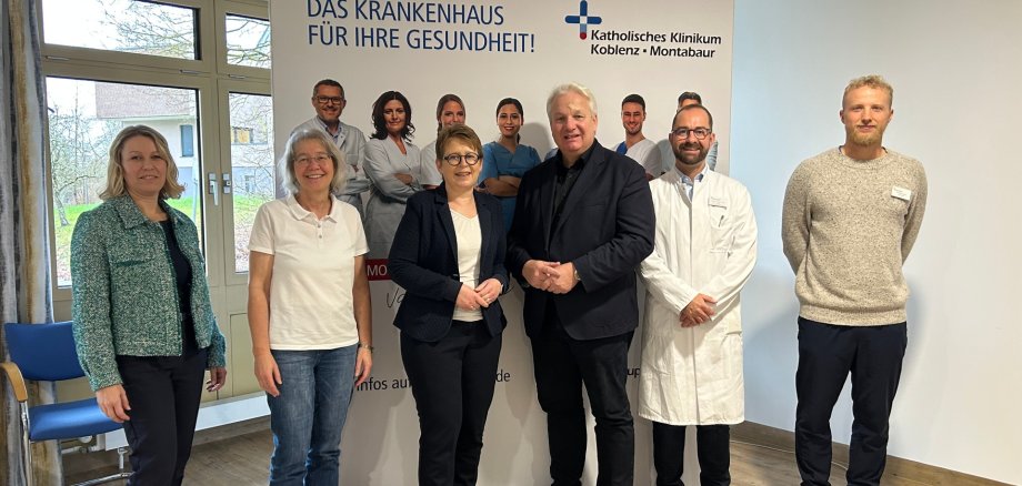 Im Bild von links nach rechts stehend in einem hellen Raum mit Holzboden und hellen Wänden, im Hintergrund ein bodenlanges Fenster, teilweise vor dem Fenster ein Roll-up mit Werbung vom Klinikum: Fr. Saal, Fr. Dr. Maxeiner, Fr. Dr. Machalet, Hr. Isenberg, Hr. Prof. Dr. Fink, Hr. Heuser