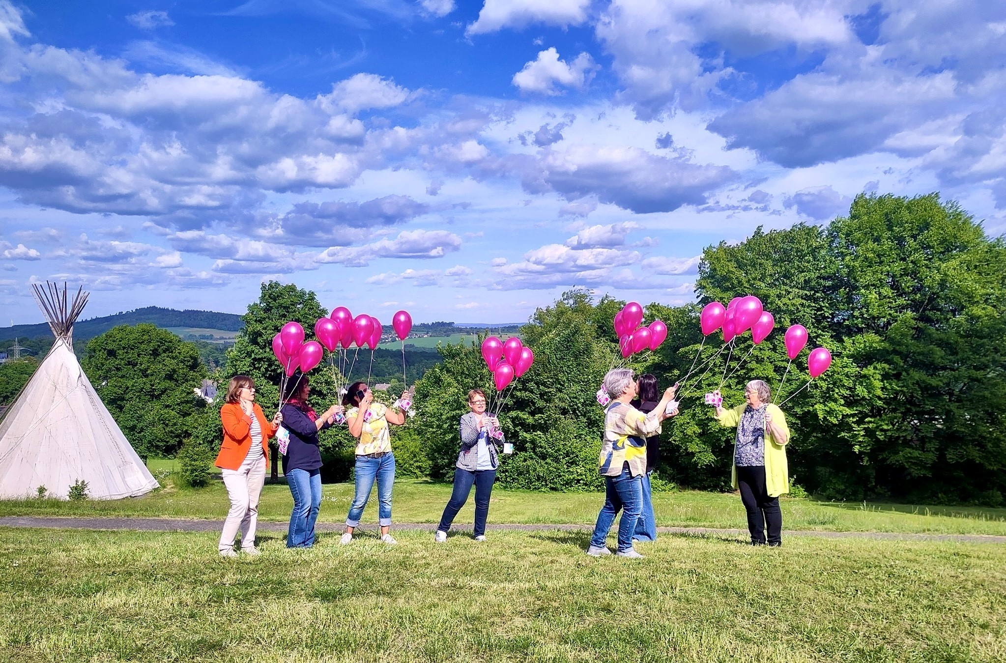 Eine Wiese, am linken Bildrand im Hintergrund ein weißes Indianer-Tipi, rechts belaubte Bäume. Mittig im Bild 7 Frauen in bunter Kleidung, zentral Dr. Tanja Machalet. Alle halten an pinkfarbene Ballons, an deren Schnüren kleine Flyer mit dem Allize-Logo und Informationen zur App befestigt sind. Der Himmel ist strahlendblau und mit einzelnen Wolken überzogen.