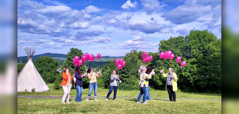 Eine Wiese, am linken Bildrand im Hintergrund ein weißes Indianer-Tipi, rechts belaubte Bäume. Mittig im Bild 7 Frauen in bunter Kleidung, zentral Dr. Tanja Machalet. Alle halten an pinkfarbene Ballons, an deren Schnüren kleine Flyer mit dem Allize-Logo und Informationen zur App befestigt sind. Der Himmel ist strahlendblau und mit einzelnen Wolken überzogen. 