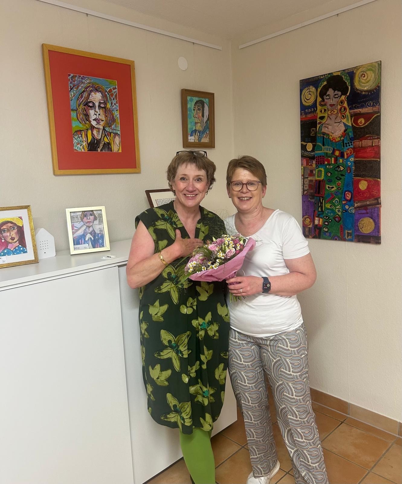 Dr. Tanja Machalet überreicht der Künstlerin Vera Apel-Jösch einen Blumenstrauß mit Wiesenblumen. Im Hintergrund sind einige Bilder zum Teil an der Wand hängend und zum Teil auf einem weißen Büroschrank aufgestellt in kleinen Rahmen zu erkennen.u sehen