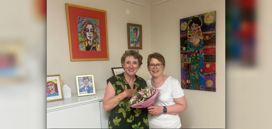 Dr. Tanja Machalet überreicht der Künstlerin Vera Apel-Jösch einen Blumenstrauß mit Wiesenblumen. Im Hintergrund sind einige Bilder zum Teil an der Wand hängend und zum Teil auf einem weißen Büroschrank aufgestellt in kleinen Rahmen zu erkennen.u sehen