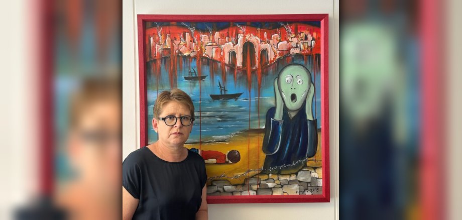 Dr. Tanja Machalet vor dem Gemälde des Nauorter Künstlers Heinz Kuppler. Das Gemälde ist an das bekannte Werk von Edvard Munch "Der Schrei" angelehnt. Im Vordergrund eine stilisierte Person, die mit weit aufgerissenem Mund zu schreien scheint, im Hintergrund in sehr kräftigen Farben eine brennende Stadt, weiter vorne das Meer mit zwei Booten, die nur aus Schatten zu bestehen scheinen. Ganz im Vordergrund ein Kinderkörper am Strand in Erinnerung an Alan Kurdi, der auf der Flucht ertrunken ist und dessen Foto um die Welt ging.