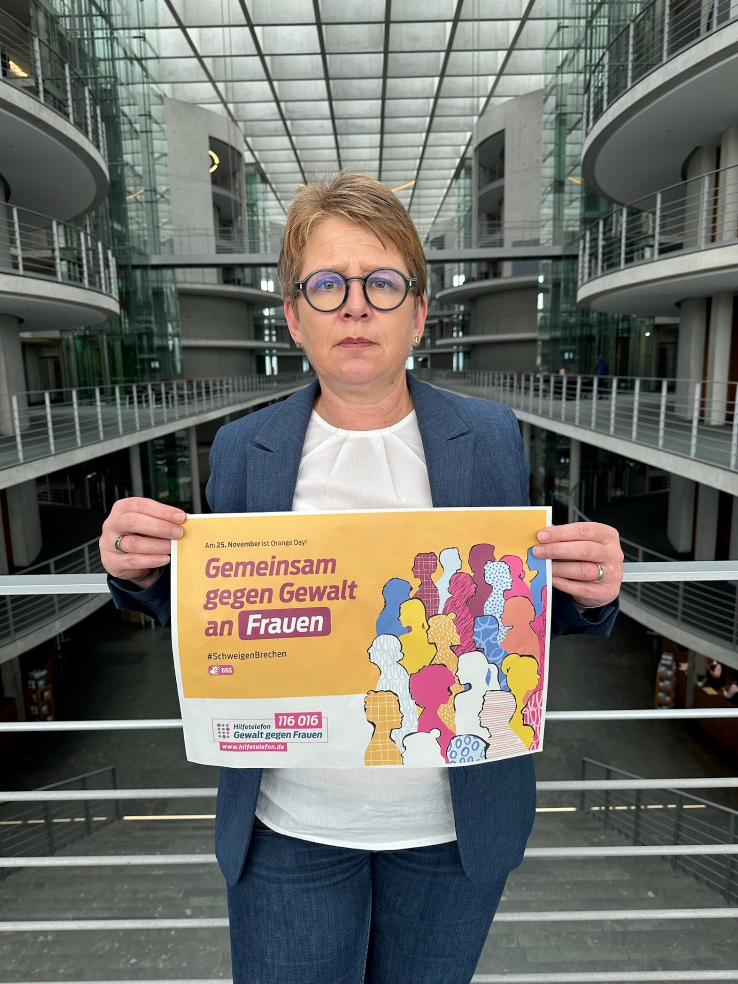 Dr. Tanja Machalet steht in einem Lichtdurchfluteten Gebäude mit Glas- und Metallarchitektur. Sie hält ein Plakat mit der Aufschrift "Gemeinsam gegen Gewalt an Frauen" sowie farbigen Silhouetten verschiedener Frauenfiguren. Das Plakat verweist auf den 25. November (Orange Day) und zeigt die Nummer des Hilfetelefons "Gewalt gegen Frauen" 116016
