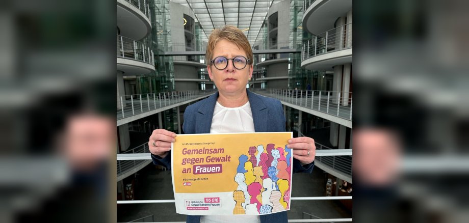 Dr. Tanja Machalet steht in einem Lichtdurchfluteten Gebäude mit Glas- und Metallarchitektur. Sie hält ein Plakat mit der Aufschrift "Gemeinsam gegen Gewalt an Frauen" sowie farbigen Silhouetten verschiedener Frauenfiguren. Das Plakat verweist auf den 25. November (Orange Day) und zeigt die Nummer des Hilfetelefons "Gewalt gegen Frauen" 116016