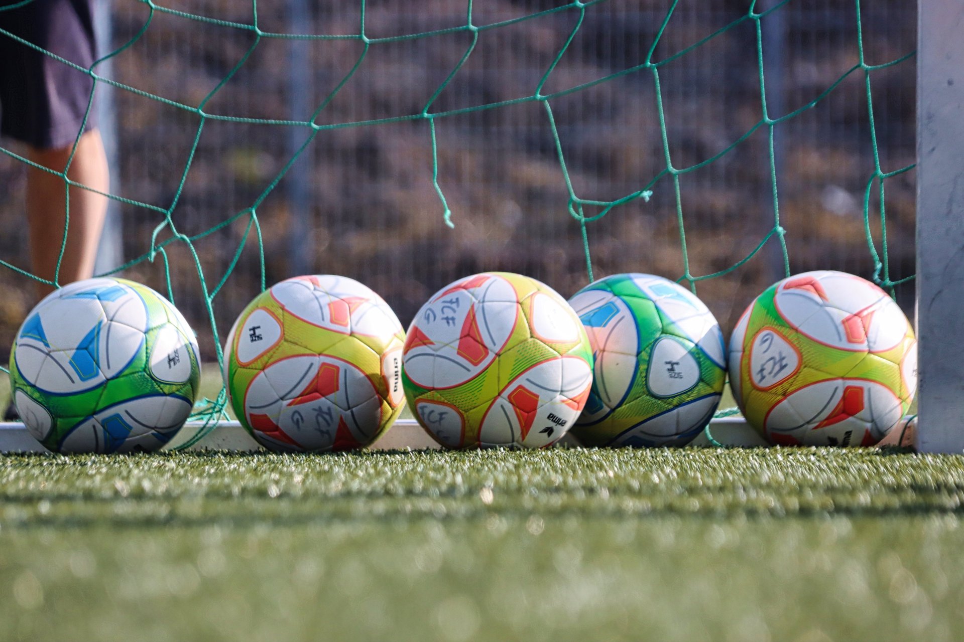 Kunstrasen auf dem fünf bunte Fußbälle vor einem Netz liegen